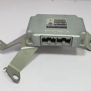 May include: A silver-colored electronic control unit with a metal bracket attached. The unit has a series of black connectors and a label with text and a barcode. The bracket has mounting holes. The item is likely an automotive part.