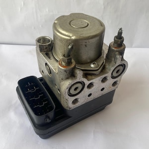 Op de afbeelding: Een zilverkleurig auto-onderdeel met een cilindrische bovenkant, rechthoekige basis en meerdere poorten en connectoren. Het onderdeel heeft een complex ontwerp met diverse schroeven en fittingen, en een zwarte elektrische connector.