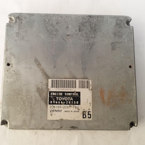 May include: A silver-colored Engine Control Module (ECM) with a label that reads "ENGINE CONTROL TOYOTA" and other technical details. The ECM has a rectangular shape with rounded corners and several mounting holes.
