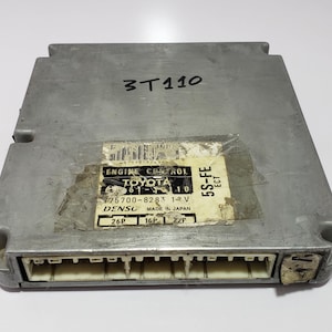 May include: A silver-colored Toyota engine control module with the text "3T110" and a label that reads "ENGINE CONTROL TOYOTA 75700-8283 19 V DENSO MADE IN JAPAN 5S-FE ECT". The module has a series of connectors.