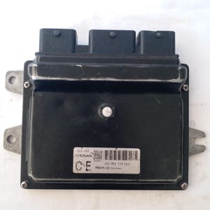 May include: A black Nissan engine control unit (ECU) with three black connectors on top. The ECU has a silver label with text and a barcode. The unit is rectangular with mounting tabs on the sides. The text "NISSAN" is visible on the label.