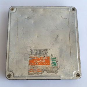 May include: A silver-colored Toyota engine control unit (ECU) with a sticker that reads "TOYOTA" and other technical information. The ECU is rectangular with rounded corners and is secured by four screws.