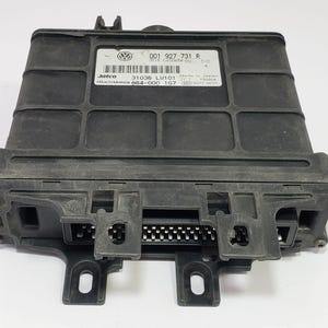 May include: A black electronic control unit with a rectangular shape. The unit has a label with text and a Volkswagen logo. It features multiple connectors and mounting brackets. The unit is likely for automotive use.