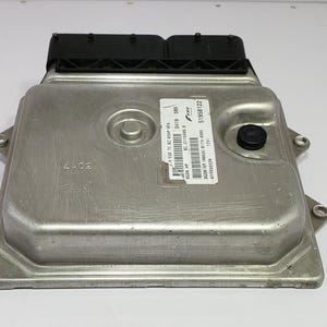 May include: A silver-coloured electronic control unit (ECU) with a black connector and a black cap. The ECU has a rectangular shape with rounded corners and several mounting holes. A label with text and barcodes is affixed to the top.