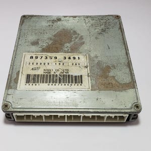 Puede incluir: Una unidad de control electrónico (ECU) rectangular de color plateado con una etiqueta que dice "Nikki Co. Ltd. Made in Japan". La ECU tiene una serie de conectores en un lado y está asegurada con cuatro tornillos. La etiqueta incluye los números "897359 3491".