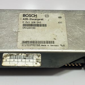 Pode incluir: Uma unidade de controle Bosch ADS-Steuergerät prateada com várias etiquetas e códigos de barras. A unidade tem um conector preto e o texto "Made in Germany" é visível. A unidade é retangular com cantos arredondados.