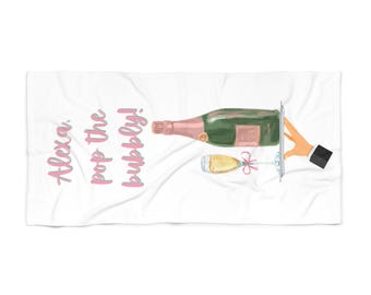 May include: White beach towel with pink text that says "Alexa, pop the bubbly!" and a cartoon illustration of a hand holding a tray with a bottle of champagne and a glass of champagne.