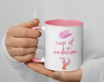 May include: White ceramic mug with a pink rim and handle. The mug has a pink cowboy hat and boots design with the text "cup of ambition".