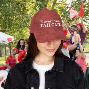 May include: A rust-colored corduroy baseball cap featuring the white embroidered text Never lost a TAILGATE across the front panel. The cap has a structured crown, a curved brim, and a fabric strap closure at the back for an adjustable fit.