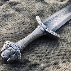 May include: Close-up of a silver sword with an intricate patterned blade. The hilt is wrapped in a textured silver material, and the guard and pommel feature detailed engravings. The sword rests on a textured, neutral-toned fabric.