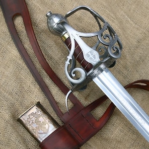 Op de afbeelding: Een gedetailleerde afbeelding van een rapierzwaard met een zilveren gevest en een bruin lederen handvat. Het lemmet is zilverkleurig en de schede is van bruin leer met een decoratieve metalen bovenkant. Het zwaard staat op een getextureerde, beige achtergrond.