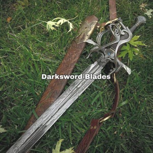 Puede incluir: Espada decorativa con una hoja estampada y una empuñadura ornamentada, sobre hierba verde junto a una vaina de cuero marrón. El texto "Darksword-Blades" es visible en la imagen.