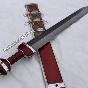 Könnte beinhalten: Ein römisches Gladius-Schwert mit dunkler Klinge und rotem Holzgriff. Der Knauf und die Scheide sind mit silbernen Akzenten und dekorativen Elementen versehen. Die Scheide ist rot mit silberner Verzierung. Das Schwert liegt auf einer weißen Oberfläche.