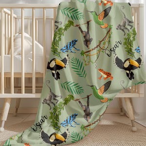 May include: A light green baby blanket with a jungle animal pattern. The blanket features toucans, monkeys, hummingbirds, and frogs. The name "Oscar" is written on the blanket in a cursive font. The blanket is draped over a white crib.