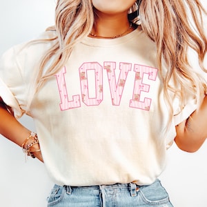 Puede incluir: Camiseta de color marfil con la palabra "LOVE" en letras grandes y rosas. Las letras tienen un patrón de rayas y están rellenas de pequeños diseños florales y de ositos de peluche. La camiseta se combina con vaqueros azules.