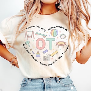 Può includere: T-shirt color avorio con un design a forma di cuore contenente le lettere "OT" e illustrazioni di attrezzature per parchi giochi. Il design include testo come "Executive Functioning" e "Sensory Processing", il tutto in una palette di colori pastello.
