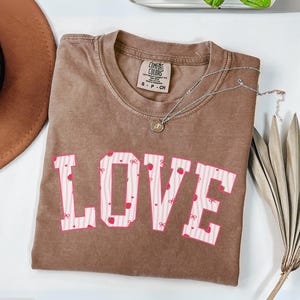 Puede incluir: Camiseta color espresso con la palabra "LOVE" en rosa, adornada con fresas y lazos. Un collar plateado con un colgante descansa sobre la camiseta. La camiseta está doblada sobre una superficie blanca.