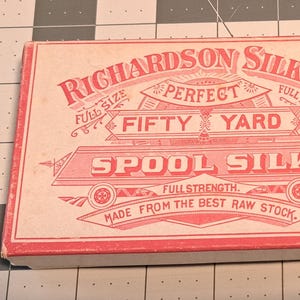 Puede incluir: Una caja rectangular de hilo de seda de la empresa Richardson. La caja es de color rosa claro con letras y gráficos rojos. El texto incluye "Perfect Fifty Yard Spool Silk" y "Made from the best raw stock."