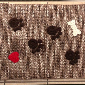 May include: A brown and white crocheted dog bed with paw prints, a bone, and a red heart.