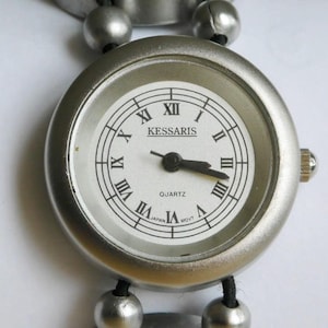 May include: A silver wristwatch with a white face and black Roman numeral hour markers. The watch face also features the text "KESSARIS QUARTZ".