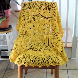 May include: A yellow crocheted doily or tablecloth draped over a wooden chair. The doily has a circular design with a central flower motif.