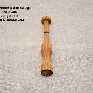 May include: A wooden Yarn Worker's Sett Gauge made of red oak. The tool is 4.5 inches long with a shaft diameter of .250 inches. The gauge has a cylindrical shape with a flared base and a small top.