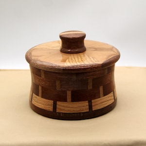 Puede incluir: Una caja de madera con tapa, hecha de diferentes tipos de madera. La caja tiene un patrón geométrico y una perilla en la tapa.