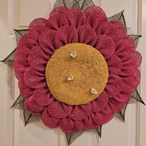 May include: A large, decorative flower wreath with a magenta mesh petal design and a yellow center. The center features three small bee embellishments. The wreath is accented with dark green mesh leaves, creating a cheerful, nature-inspired home decor item.