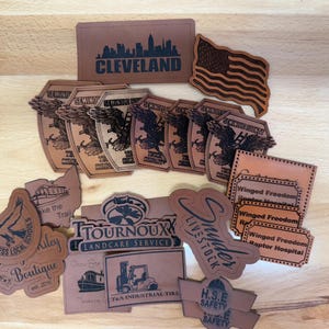 May include: A collection of tan faux leather patches featuring various laser-engraved designs, including the Cleveland skyline, an American flag, Seminole County Truck Company logos, Winged Freedom Raptor Hospital labels, and business logos.