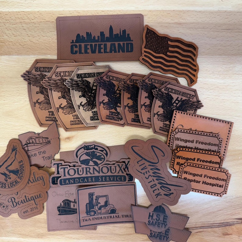 May include: A collection of tan faux leather patches featuring various laser-engraved designs, including the Cleveland skyline, an American flag, Seminole County Truck Company logos, Winged Freedom Raptor Hospital labels, and business logos.