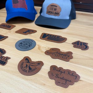 May include: Two baseball caps, one blue with an American flag patch and one grey and black with an IBEW Local 540 patch, are displayed alongside various custom-shaped leather and grey patches featuring diverse logos, text, and graphic designs.