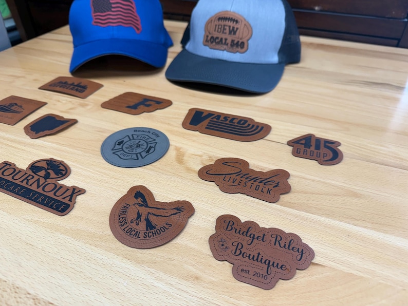 May include: Two baseball caps, one blue with an American flag patch and one grey and black with an IBEW Local 540 patch, are displayed alongside various custom-shaped leather and grey patches featuring diverse logos, text, and graphic designs.