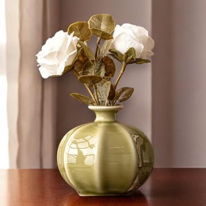 May include: A green ceramic vase with a rounded shape and a glossy finish. The vase contains three white rose blossoms with green leaves. The vase is on a wooden surface.