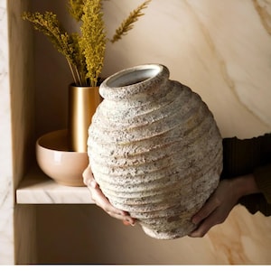 Könnte beinhalten: Eine große, weiße, strukturierte Keramikvase mit rundem Korpus und einer breiten Öffnung. Die Vase hat ein rustikales, gealtertes Aussehen und ist perfekt für die Präsentation von Trockenblumen oder Zweigen.