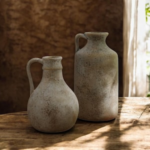 Op de afbeelding: Twee beige keramische kruiken met handvatten. De kruiken hebben een getextureerd, gespikkeld oppervlak en een rustieke uitstraling. Eén kruik is korter en ronder, de andere is hoger en cilindrischer. Ze staan op een houten oppervlak.