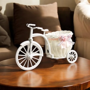 May include: A white decorative tricycle with a woven basket. The basket is adorned with a pink bow. The tricycle has a classic design with spoked wheels and a seat. The decorative piece is on a wooden table.