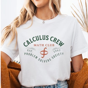 May include: A white short-sleeved t-shirt featuring a graphic design with the text CALCULUS CREW in dark green, MATH CLUB in orange, and EST. 1982 PROBLEM SOLVERS SOCIETY in dark green, centered around an orange integral symbol graphic.