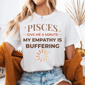 Puede incluir: Camiseta blanca con el texto "PISCES - GIVE ME A MINUTE - MY EMPATHY IS BUFFERING" en letras marrones. La camiseta presenta un gráfico de un símbolo de carga en naranja y amarillo. La modelo lleva pantalones cortos vaqueros y una rebeca color mostaza.