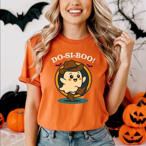 May include: Orange t-shirt with the text "DO-SI-BOO!" above a cartoon ghost wearing a cowboy hat and boots. The shirt is part of a Halloween theme, with pumpkins and bats in the background.