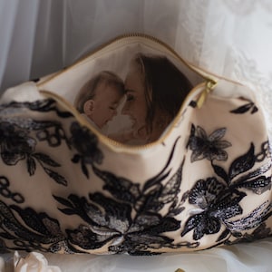 May include: A beige zippered pouch with black floral embroidery. The pouch is open and a photo of a mother and child is visible inside.