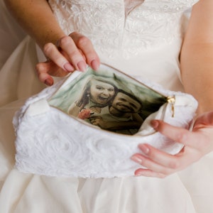 May include: A white, fluffy clutch bag with a zipper closure. The bag is open, revealing a photo of a couple inside. The photo shows a woman with long, dark hair and a man with short, dark hair. They are both smiling.