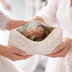 May include: A cream-colored, beaded cosmetic bag with a gold zipper, held open to reveal a printed photograph of two women. The bag is embellished with intricate beadwork and held by two people.