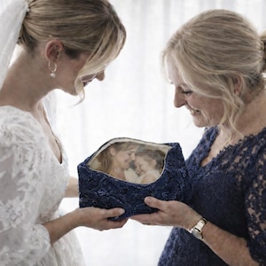 May include: A bride in a white lace wedding dress and veil and another woman in a navy blue lace dress are looking at a navy blue box with a photo inside. The box is held between them.