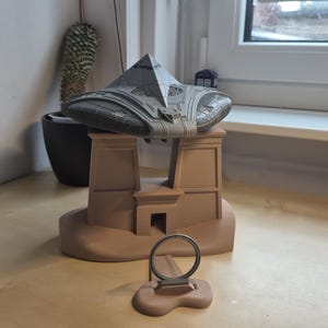Puede incluir: Una escultura gris de aspecto futurista de una nave espacial descansa sobre un modelo arquitectónico de color beige. Un pequeño objeto circular se encuentra frente al modelo. Un cactus en una maceta negra es visible en el fondo.