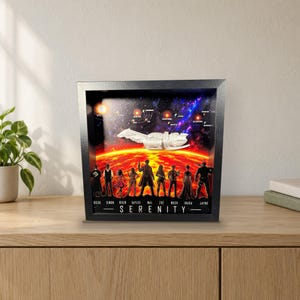 Könnte beinhalten: Ein schwarzer Schattenfugenrahmen mit einem 3D-Diorama des Raumschiffs Serenity über einem roten Planeten. Unter dem Schiff befinden sich Silhouetten von neun Charakteren mit ihren Namen und das Wort SERENITY in der unteren Mitte gedruckt.