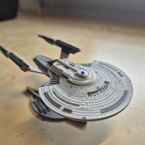 Könnte beinhalten: Ein detailliertes Modell eines Raumschiffs mit einem scheibenförmigen Hauptrumpf und zwei Warpgondeln. Das Modell ist hauptsächlich grau mit schwarzen und roten Akzenten, und der Text "NCC-1701-B" ist sichtbar. Das Modell steht auf einem runden Sockel.