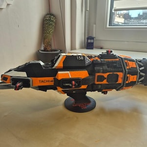 Könnte beinhalten: Ein detailliertes Modell des Tachi-Raumschiffs in Schwarz und Orange mit der aufgedruckten Nummer 158 an der Seite. Das Raumschiff ist auf einem runden schwarzen Ständer mit Text auf der Basis montiert und zeigt komplexe mechanische Details sowie ein großes hinteres Triebwerk.
