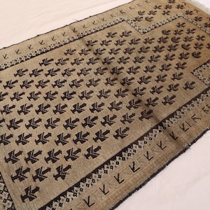 May include: A rectangular rug with a beige background and a repeating dark brown pattern. The rug has a border with geometric designs and fringe on the edges. The rug's dimensions are not visible.