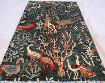 May include: A rectangular, hand-knotted rug with a dark teal background. The rug features a detailed pattern of trees, birds, and floral elements in shades of red, orange, white, and blue. The rug has a fringe along the bottom edge.