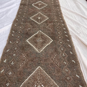 May include: A brown runner rug with a geometric diamond pattern in white. The rug has a textured surface and is rectangular in shape. The design features repeating diamond shapes and small white dots.